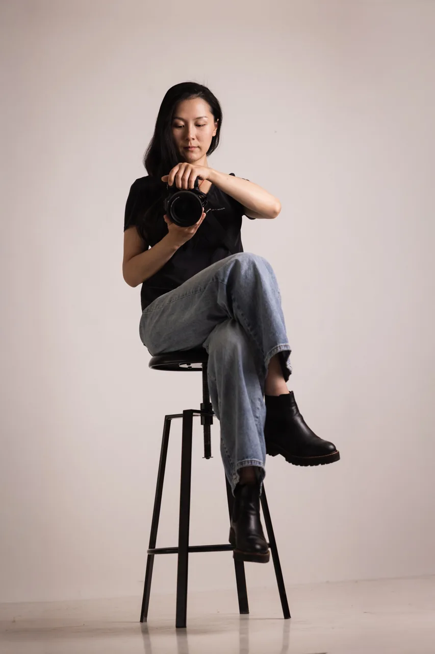 A photographer seated on a stool holding a camera against a warm studio backdrop